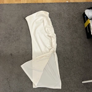 Vit långklänning från SHEIN - Snygg vit långklänning från SHEIN i storlek S. Klänningen är axelbandslös med en draperad detalj på sidan och har en slits framtill. Materialet är mjukt och har en lätt glansig finish, perfekt för dig som vill sticka ut med en clean och elegant look.
