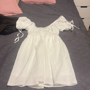 Vit kort klänning med puffärmar - Superfin vit kort klänning från Stradivarius med puffiga kortärmar och knytband vid axlarna. Klänningen har en smockad midja och är gjord i ett strukturerat, lätt tyg som känns luftigt. Perfekt för varma dagar och ger en söt, romantisk vibe.