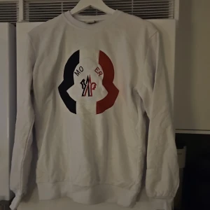 Vit sweatshirt - Säljer en vit sweatshirt i storlek L. Tröjan har ett stort broderat logo på bröstet i svart, vitt och rött. Klassisk rund halsringning och ribbade muddar vid ärmslut och nederkant. Perfekt för dig som gillar stilrena märkesplagg. Passar även M. Helt ny!!!