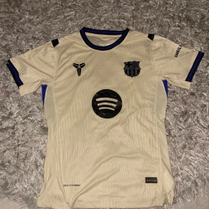 Barcelona Ferran 7 bortatröja Nike - Snygg beige Barcelona bortatröja med svarta detaljer, Ferran Torres nummer 7 på ryggen och Spotify-logga på bröstet. Tröjan har korta ärmar, blå och svarta inslag vid ärmslut och sidor, samt Dri-FIT material för extra komfort.
