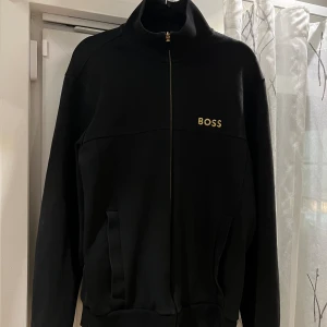 Svart ziptröja från BOSS - Tja! säljer min Boss ziptröja, då jag inte har någon användning av den längre. Har använt den fåtals gånger. Det är en svart långärmad ziptröja från BOSS med guldfärgad logga på bröstet. Tröjan har hög krage, hel dragkedja och två fickor framtill. Perfekt för en clean och stilren look. 