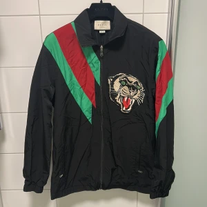 Gucci svart vindjacka med pantermärke storlek M - Svart vindjacka från Gucci med gröna och röda diagonala ränder över axlarna och ett stort broderat pantermärke på bröstet. Jackan har dragkedja framtill, hög krage och elastiska muddar. Materialet är lätt och glansigt, perfekt för dig som vill sticka ut. 