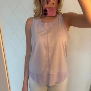 Ljuslila träningslinne från Adidas - Säljer en ljuslila träningslinne från Adidas med brottarrygg och diskret logga i nacken. Toppen är ärmlös och har en lös passform, perfekt för träning eller varma dagar. Tillverkad i ett lätt och svalt syntetmaterial som andas.