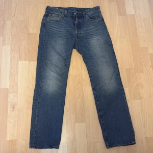 Levi's 501 blå jeans straight fit - Klassiska blå Levi's 501 jeans med rak passform och knappgylf. Jeansen har fem fickor, subtila slitningar och den ikoniska läderpatchen bak i midjan. Perfekta för dig som gillar tidlös denimstil. Storlek: W 32 L 32.