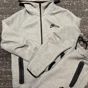 Nike tech tracksuit - Säljer en ljusgrå Nike hoodie i storlek S med hel dragkedja framtill, svarta detaljer och huva. Tröjan har långa ärmar, praktiska fickor med dragkedja och justerbar nederkant. Perfekt för chill eller träning.