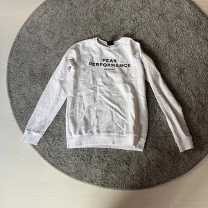 Vit Peak Performance sweatshirt - Snygg vit sweatshirt från Peak Performance med svart textprint på bröstet. Tröjan har rund hals, långa ärmar och ribbade muddar vid ärmslut och nederkant. Perfekt för dig som gillar stilren och sportig look. Storlek 170 men passar mig som har S.