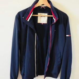 Marinblå vindjacka Tommy Jeans M - Snygg marinblå vindjacka från Tommy Jeans med klassiska röda, vita och blå detaljer på krage och ärmslut. Jackan har dragkedja, ribbade muddar och diskret logga på ärmen. Perfekt för dig som gillar sportig och stilren look.