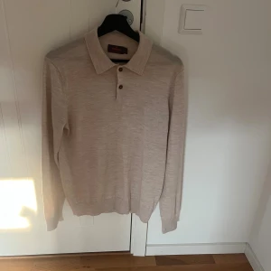 Beige merinoull pikétröja Morris M - Snygg beige pikétröja från Morris i 100% merinoull. Tröjan har klassisk krage, knäppning med två knappar och lång ärm. Perfekt för dig som gillar stilrena och bekväma plagg med en lyxig känsla. Nypriset på denna ligger på 1700
