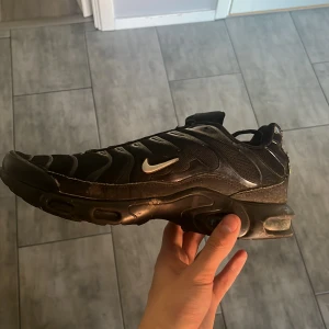 Svarta Nike Air Max Tn  - Nike Air Max Plus sneakers i svart med silvriga detaljer och TN-logga på hälen. Skon har en chunky sula med synliga luftkuddar, snörning och meshpartier för extra ventilation. Snyggt vågigt mönster på ovansidan och klassisk Nike Swoosh på sidan.