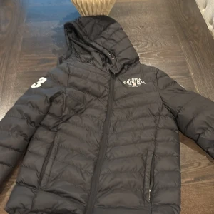 Svart pufferjacka Vinson Original - Svart pufferjacka från Vinson Original med huva och dragkedja. Jackan har vita tryck med siffran 23 på ärmen och logga på bröstet. Quiltad design och två sidofickor med dragkedja. Perfekt för kalla dagar och snygg streetstil.