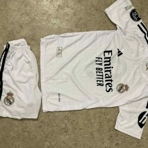 Real Madrid Mbappé fotbollströja set - Säljer ett Real Madrid fotbollströja och shorts set från Adidas, med Mbappé och nummer 9 på ryggen. Tröjan är vit med svarta detaljer, klubbmärke på bröstet och sponsortryck. Materialet är lätt och andas, perfekt för träning eller match.
