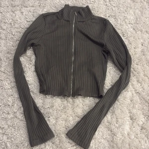 Ribbad grå kofta från H&M Divided XS - Snygg croppad kofta i mörkgrått från H&M Divided, storlek XS. Tröjan har ribbad struktur, lång ärm och dragkedja framtill. Perfekt att slänga på över en topp för en chill look. Materialet är mjukt och stretchigt, vilket gör den skön att bära.