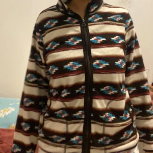 Mönstrad fleecejacka med dragkedja - Fleecejacka med brunt, beige och blått mönster, hel dragkedja och hög krage. Jackan har ett coolt rutigt och etno-inspirerat mönster, två fickor framtill och är långärmad. Står strl 14 men tycker den är stor för mig som är XS 🤍