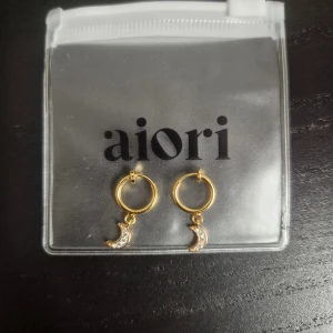Guldiga örhängen med månberlocker från Aiori - Söta guldiga örhängen för folk utan piercings från Aiori med små månar som hänger och är dekorerade med glittrande stenar. För tunnare öronlob.