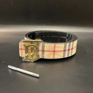 Svart bälte med guldspänne - Säljer ett bälte med ikoniskt rutigt mönster i beige, svart och rött samt ett elegant guldigt B-spänne. Bältet har två sidor: en klassisk beige-rutig och en svart med diskret reliefmönster. Perfekt accessoar för att lyfta din outfit.