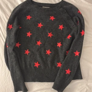 Grå stickad tröja med röda stjärnor Zadig & Voltaire - Mörkgrå stickad tröja från Zadig & Voltaire med röda stjärnor över hela tröjan. Rund halsringning och dekorativa knappar på ena axeln. Långärmad och i mjukt material som passar perfekt till höst och vinter.