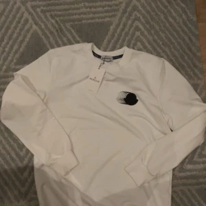 Vit långärmad tröja från Moncler - Snygg vit långärmad tröja från Moncler i bomull. Tröjan har en svart Moncler-logga med coolt grafiskt tryck på bröstet och rund halsringning. Perfekt för dig som gillar stilrena och trendiga plagg med exklusiv känsla.