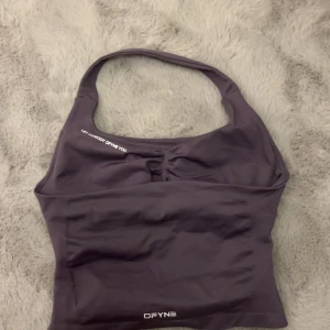 Lila träningstopp från DFYNE - Säljer en lila/grå färgad halterneck träningstopp från DFYNE.  Toppen är croppad och har en snygg rynkdetalj framtill. Materialet är stretchigt och består av 90% nylon och 10% elastan. Färgen heter Truffle på hemsidan.