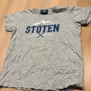 Grå t-shirt från Stöten Sälen - Snygg grå t-shirt från Stöten Sälen Sweden med blå och vit logga framtill. Klassisk passform med rund hals och korta ärmar. Perfekt för dig som gillar enkel och sportig stil. Tillverkad i mjuk bomull för skön känsla hela dagen.