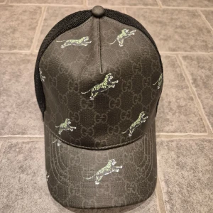 Gucci svart keps med tigerprint - Svart keps från Gucci med mesh på baksidan och justerbar rem. Framsidan har GG-monogram och gröna tigertryck över hela kepsen. Snygg och unik design som sticker ut, perfekt för dig som gillar streetstyle och exklusiva märken. Storlek M 57 cm, ställbar i storleken.