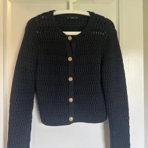 Mörkblå stickad kofta från MNG - Säljer en mörkblå stickad kofta från MNG med snyggt hålmönster och guldiga knappar framtill. Koftan har rund halsringning och långa ärmar, perfekt att slänga över en topp för en chill look.