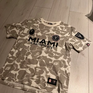 Miami fotbollströja BAPE x A Bathing Ape - Unik fotbollströja från BAPE x Miami med beige och vit camouflage-mönster, svart tryck med MIAMI och BAPE 93 på ryggen. Patchar på ärmarna och bröstet, rund hals och korta ärmar. Tillverkad i mjuk bomull, perfekt för dig som gillar streetwear och fotboll.