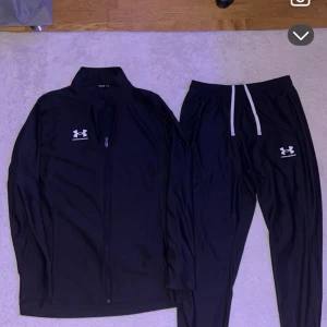 Svart träningsset från Under Armour - Snyggt svart träningsset från Under Armour med zip-tröja och matchande byxor. Tröjan har dragkedja framtill och logga på bröstet, byxorna har resår och vit snörning samt logga på benet. Perfekt för gymmet eller löprundan.