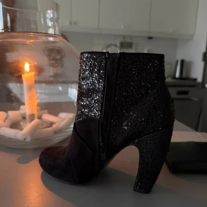 Svarta glittriga stövletter från Villain - Säljer ett par svarta stövletter från Villain med glittrig ovandel och mockaliknande material. Skorna har hög klack, rund tå och dragkedja på sidan. Perfekta för att sticka ut med en extra touch av glitter.