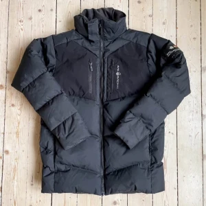 Svart pufferjacka från Sail Racing - Svart pufferjacka från Sail Racing med hög krage och flera praktiska dragkedjefickor framtill. Jackan har Sail Racing-logga på bröstet och patch på ärmen med 'Antarctica Expedition'. Materialet är syntet och dun, perfekt för kalla dagar.