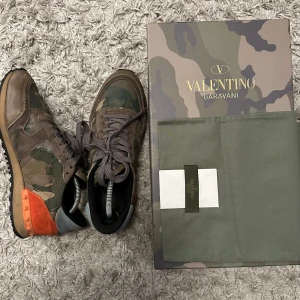 Valentino Rockrunner sneakers camouflage - Säljer ett par Valentino Rockrunner sneakers med camouflagemönster i grönt, beige och brunt. Skorna har detaljer i mocka och skinn, samt en orange häl med nitar. Endast skorna medföljer Klassisk snörning och platt sula. Perfekt för dig som gillar exklusiva streetwear-vibes. Strl41