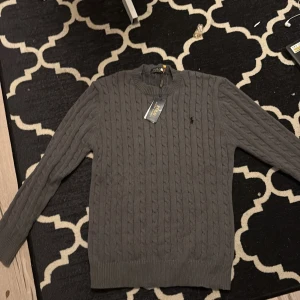 Grå kabelstickad tröja Polo Ralph Lauren - Snygg grå kabelstickad tröja från Polo Ralph Lauren med klassisk broderad logga på bröstet. Tröjan har rund halsringning, långa ärmar och ribbade muddar. Perfekt för lager-på-lager och en clean look.