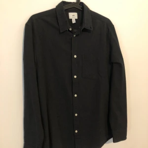 Svart skjorta från H&M Regular Fit - Svart skjorta från H&M i regular fit med klassisk krage och vita knappar framtill. Skjortan har en bröstficka och långa ärmar. Materialet är mjukt och känns som bomull, perfekt för en clean och enkel look.