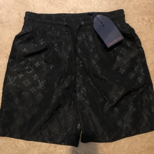 Svarta Louis Vuitton shorts med monogram - Snygga svarta shorts från Louis Vuitton med klassiskt monogrammönster över hela tyget. De har elastisk midja med snörning och innerfoder i mesh. Perfekta för dig som vill ha en lyxig och trendig look i sommar.