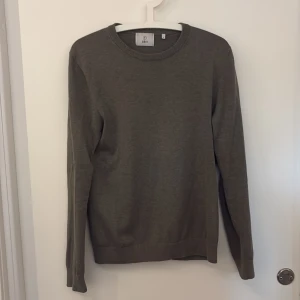 Grå-grön stickad tröja från Bläck - En stilren grå-grön stickad tröja från Bläck i storlek S. Tröjan har rund halsringning, ribbade muddar och långärmad passform. Perfekt för dig som gillar enkel och clean look. Materialet är mjukt och skönt mot huden.