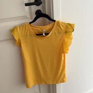 Gul ribbad topp med volangärmar från JDY - Säljer en ljusgul ribbad topp från JDY i storlek Large. Toppen har korta ärmar med snygga volangdetaljer som ger en extra touch. Materialet är mjukt och lätt, perfekt för varma dagar. Rund halsringning och croppad passform gör den superfräsch och trendig.