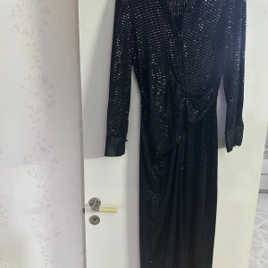 Svart glittrig långklänning från Zara - Svart långklänning från Zara med glittriga paljetter och snygg krage i satin. Klänningen har långärmade ärmar med breda manschetter och ett omlottliv som ger en cool siluett. Perfekt för dig som vill sticka ut med en glam look.