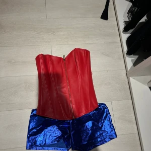 Röd korsett med snörning och dragkedja - Säljer en snygg röd korsett i syntetmaterial med dragkedja framtill och snörning baktill. Korsetten har en stilren form med markerade sömmar och är axelbandslös. Perfekt för dig som vill sticka ut på festen eller cosplay.