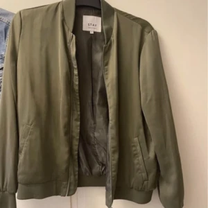 Olivgrön bomberjacka från Stay - Snygg olivgrön bomberjacka från Stay med klassisk ribbad krage och muddar. Jackan har två fickor framtill och stängs med dragkedja. Perfekt för dig som gillar en stilren och enkel look.