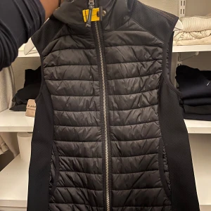 Svart dunväst från Parajumpers XS - Svart dunväst från Parajumpers i storlek XS. Västen har quiltad framsida, dragkedja hela vägen, hög krage och en gul detalj vid halsen. Sidorna är i stretchigt material för extra komfort. Perfekt för dig som gillar stila upp dig. Köp innan någon annan köper🤭