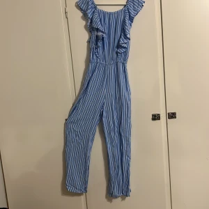 Blåvit randig jumpsuit från Kappahl - Supersöt blå och vit randig jumpsuit från Kappahl med volangdetaljer längs axelbanden och ärmlös design. Perfekt för sommarens varma dagar, med bekväm resår i midjan och mjukt, lätt material.