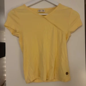 Gul ribbad topp från Ü - Snygg gul ribbad topp från Ü med korta ärmar och v-ringad omlott-design. Toppen har en liten metallbricka nertill som detalj och är gjord i mjuk bomull. Perfekt för dig som gillar färg och enkel stil.