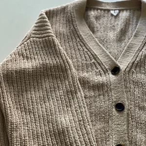 Beige stickad kofta från Arket - Säljer en beige stickad kofta från Arket i storlek L. Koftan har grovstickad struktur, v-ringad hals och svarta knappar framtill. Materialet är mjuk bomull och passformen är loose med långa ärmar. Perfekt för lager-på-lager under kyliga dagar.