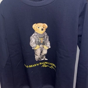 Mörkblå Polo Bear tröja Ralph Lauren - Mörkblå sweatshirt från Ralph Lauren med ikoniska Polo Bear-trycket framtill. Tröjan har rund halsringning och ribbade muddar. Perfekt för dig som vill ha en klassisk och stilren look med en lekfull twist.
