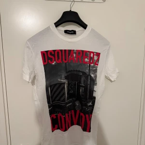 Vit DSQUARED2 t-shirt med tryck - Vit t-shirt från DSQUARED2 med stort svart och rött tryck framtill som visar en lastbil och texten 'CONVOY'. Klassisk rund hals och korta ärmar. Tillverkad i mjuk bomull, perfekt för en cool och avslappnad stil.