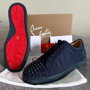 Christian Louboutin skor - Säljer ett par mörkblå sneakers från Christian Louboutin med ikonisk röd sula och svarta nitar på tån. Skorna är tillverkade i mocka med snörning och har en rund tå. Insidan är i beige skinn och yttersulan är platt. Perfekt för dig som gillar edgy designerstil.