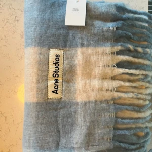 Blågrå rutig halsduk Acne Studios - Säljer en stor och fluffig halsduk från Acne Studios i en mjuk blandning av blågrå och beige rutor. Halsduken har fransar i kanterna och en tydlig Acne Studios-logga. Perfekt för att ge din outfit en cool touch under kyliga dagar. Hör av dig vid minsta fråga!
