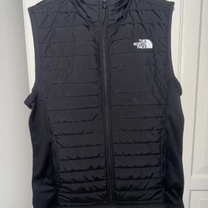 Svart dunväst från The North Face - Svart dunväst från The North Face med quiltad framsida och dragkedja hela vägen. Västen har ståkrage, två sidofickor med dragkedja och logga i vitt på bröstet och ryggen. Perfekt för lager-på-lager och outdoor. Snygg, enkel och funktionell design. Köpte den i september för 1999kr har knappt använt den! Riktigt fin väst! Mitt pris : 699