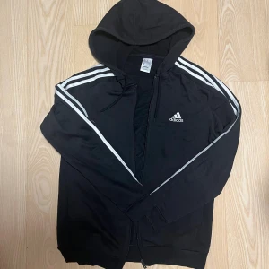 Svart Adidas hoodie med vita ränder - Svart hoodie från Adidas med klassiska vita ränder längs ärmarna och Adidas-logga på bröstet. Tröjan har huva med snörning och dragkedja framtill. Perfekt för chill eller träning, och tillverkad i mjuk polyester.
