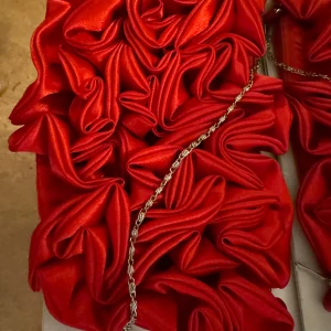 Röd axelväska med volangdetaljer - Säljer en riktigt snygg röd axelväska med coola volangdetaljer på framsidan. Väskan har en kedja som axelrem och är gjord i glansigt satinliknande material. Stängs med dragkedja och har en unik, iögonfallande design som verkligen sticker ut.