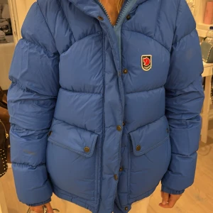 Blå pufferjacka från Fjällräven 🩵🌟 - Säljer en blå pufferjacka från Fjällräven med ikonisk röd logga på bröstet. Jackan har stora fickor med knappar, hög krage och är quiltad för extra värme. Perfekt för kalla dagar och har en snygg, lite oversized passform.